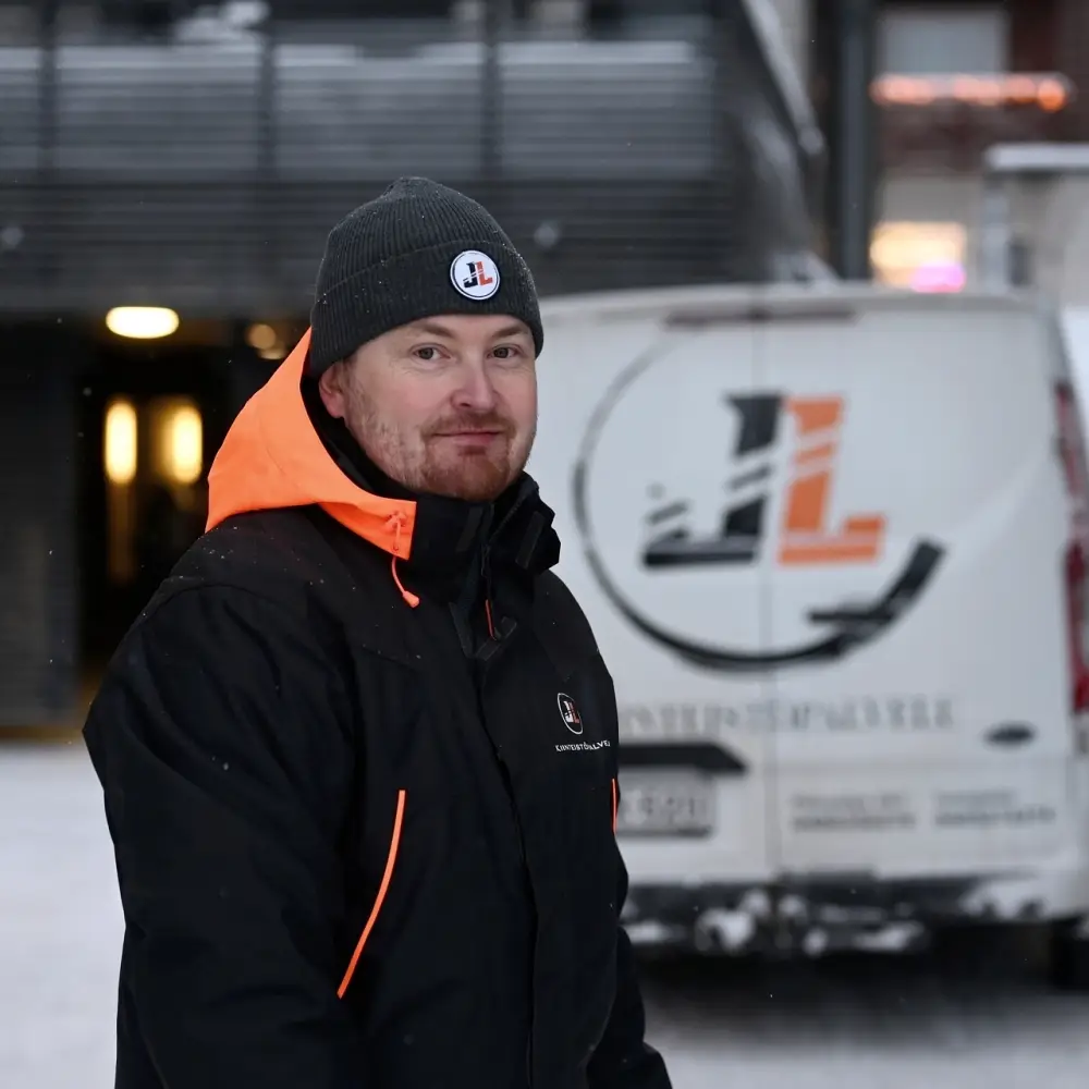 Mies talvivaatteissa seisoo lumisessa ympäristössä, taustalla kiinteistöpalveluita tarjoavan yrityksen pakettiauto.