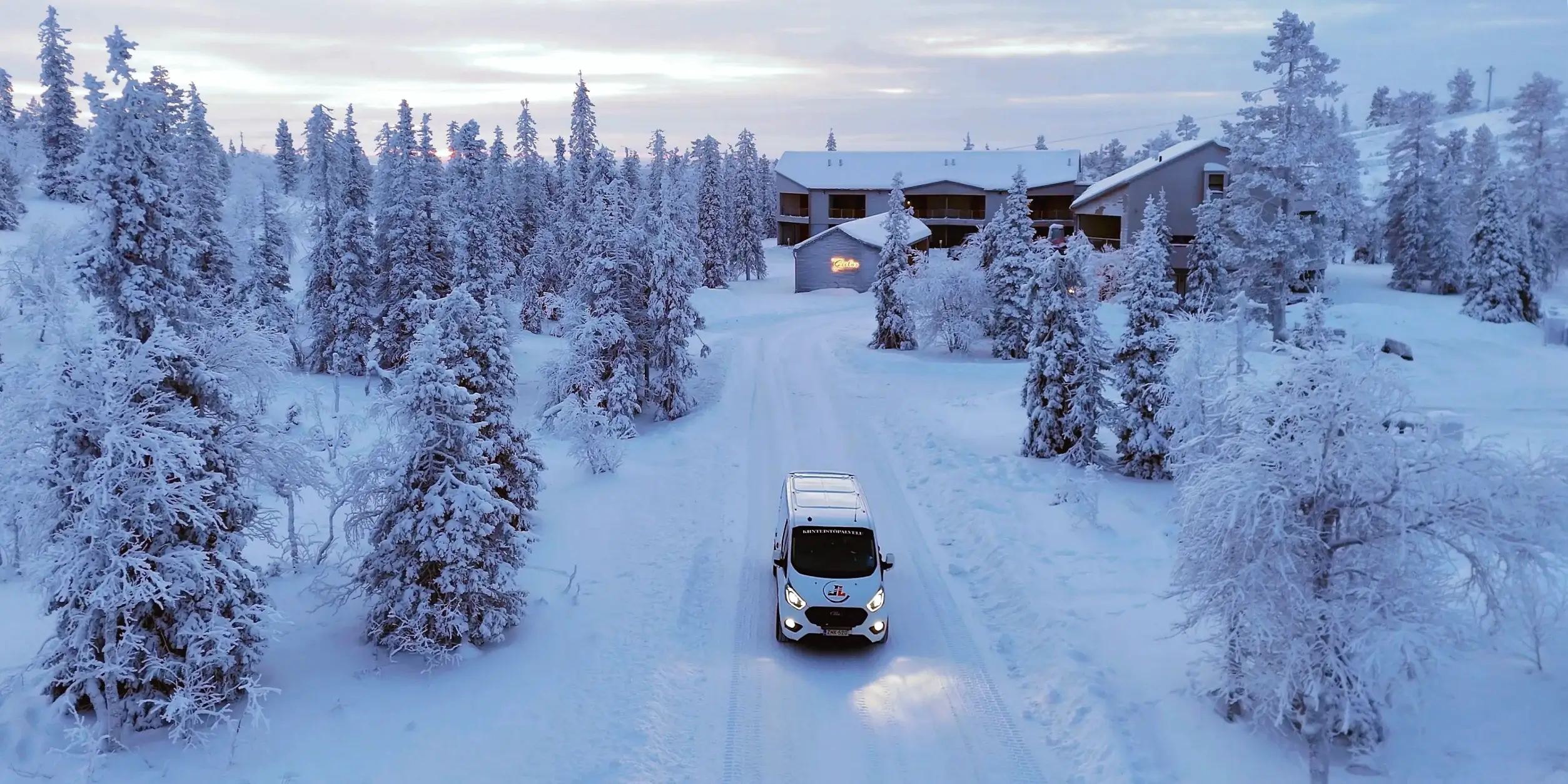 Valkoinen pakettiauto ajaa lumisella tiellä kohti rakennuksia talvisessa metsämaisemassa, jossa kiinteistöpalveluita tarvitaan.